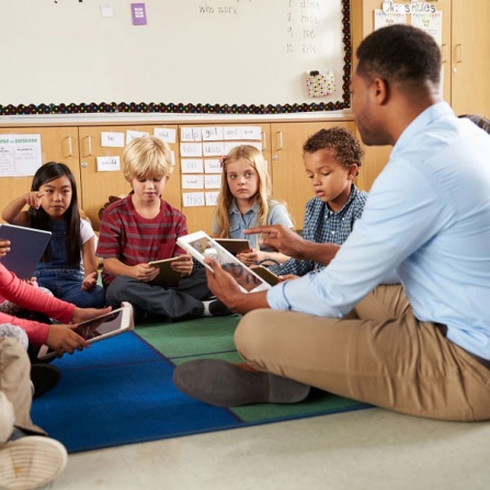 The new Center for Early Literacy and Responsible AI will focus on harnessing the power of artificial intelligence to transform early literacy instruction for culturally and linguistically diverse learners in kindergarten through second grade. Image of teacher and students sitting on floor in circle reading. Teacher and students are holding touch pads for the learning experience. 