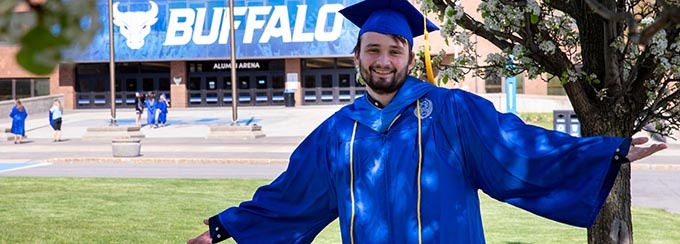 Student in graduation gown and cap in front of Alumni Arena. 