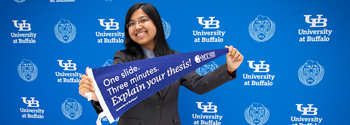 Sandipa Bhattacharjee smiling and holding her Three Minute Thesis pennant that reads: One slide. Three minutes. Explain your thesis! 