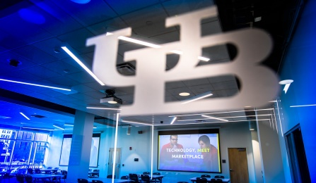 Image of the interior of UB's Incubator @ CBLS. Photo looks through glass window with UB lockup into room with tables, chairs and large screen that says, "Technology Meet Marketplace.". 